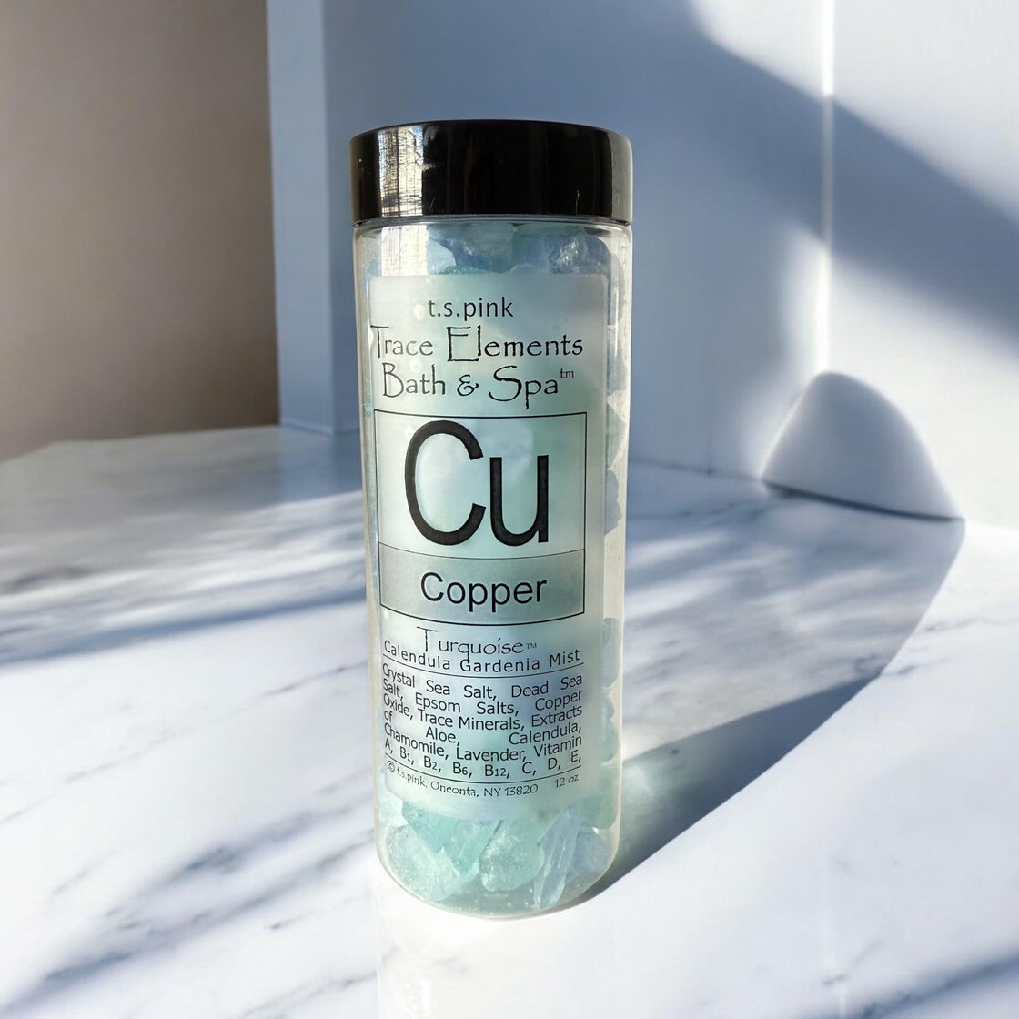 A clear plastic jar with a black lid, filled with light blue Rengöra bath salts, sits on a marble surface with soft shadows.