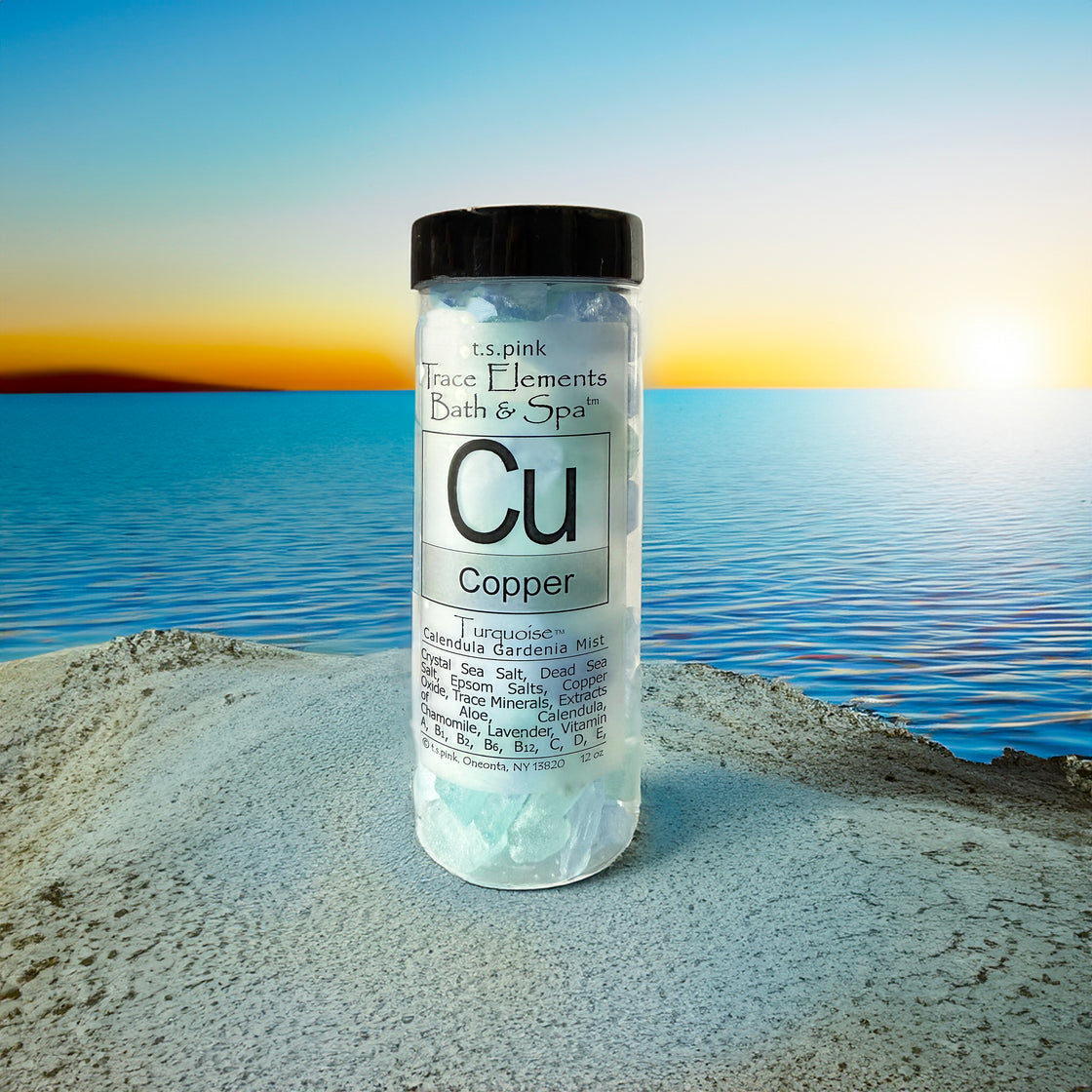 A clear plastic jar with a black lid, filled with Rengöra copper-infused bath salts, sits on a sandy surface with a tranquil ocean and sunset in the background.