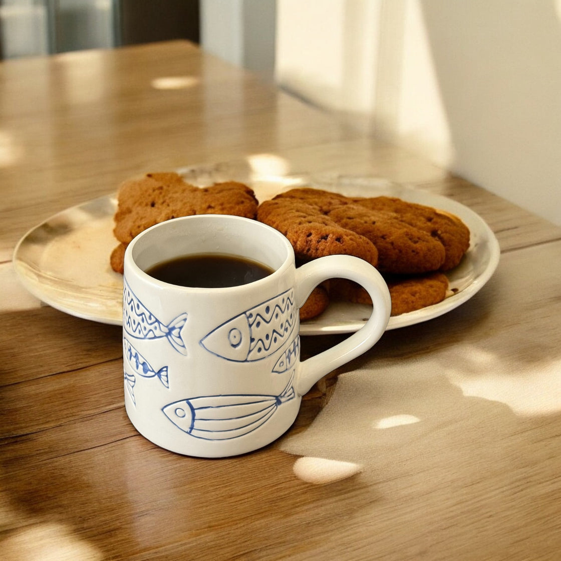 Ceramic fish mug filled with coffee placed on a wooden table, accompanied by cookies.
