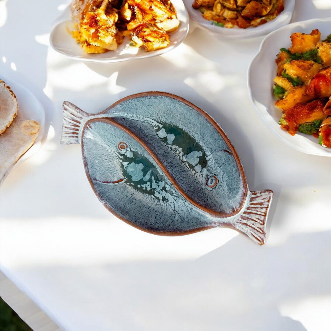 Beautifully glazed ceramic dual fish-shaped serving bowl on an outdoor table, surrounded by delicious food.