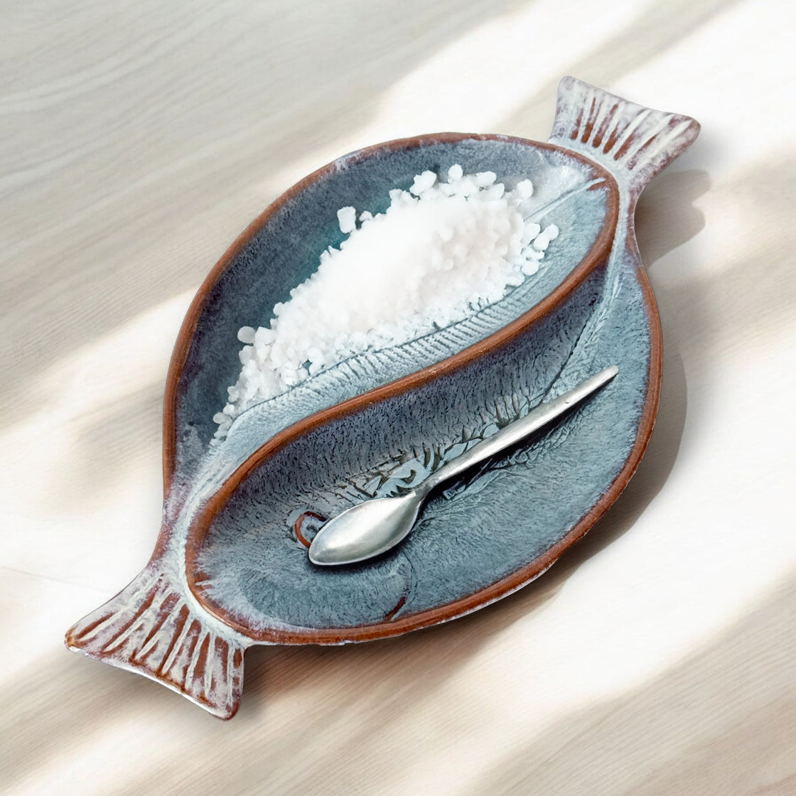 Dual fish-shaped ceramic serving bowl holding sea salt in one compartment with a small silver spoon placed elegantly inside.