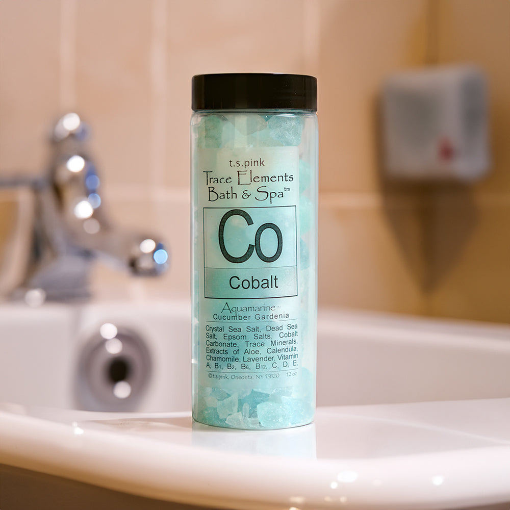 Cobalt Cucumber + Gardenia fragranced bath salts pictured in their clear cylindrical container sitting on the edge of a white bathtub.