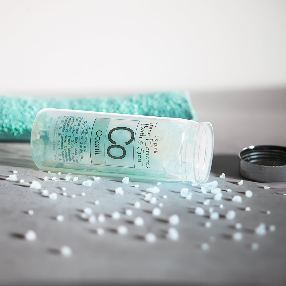 Cobalt bath crystals spread out in front of the container (laying on its side) with a teal bath towel in the background