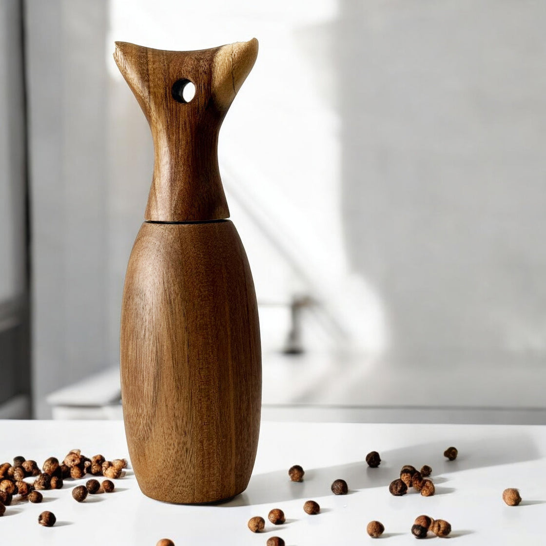 Elegant wooden pepper grinder on a bright white tabletop, surrounded by scattered peppercorns in natural light.