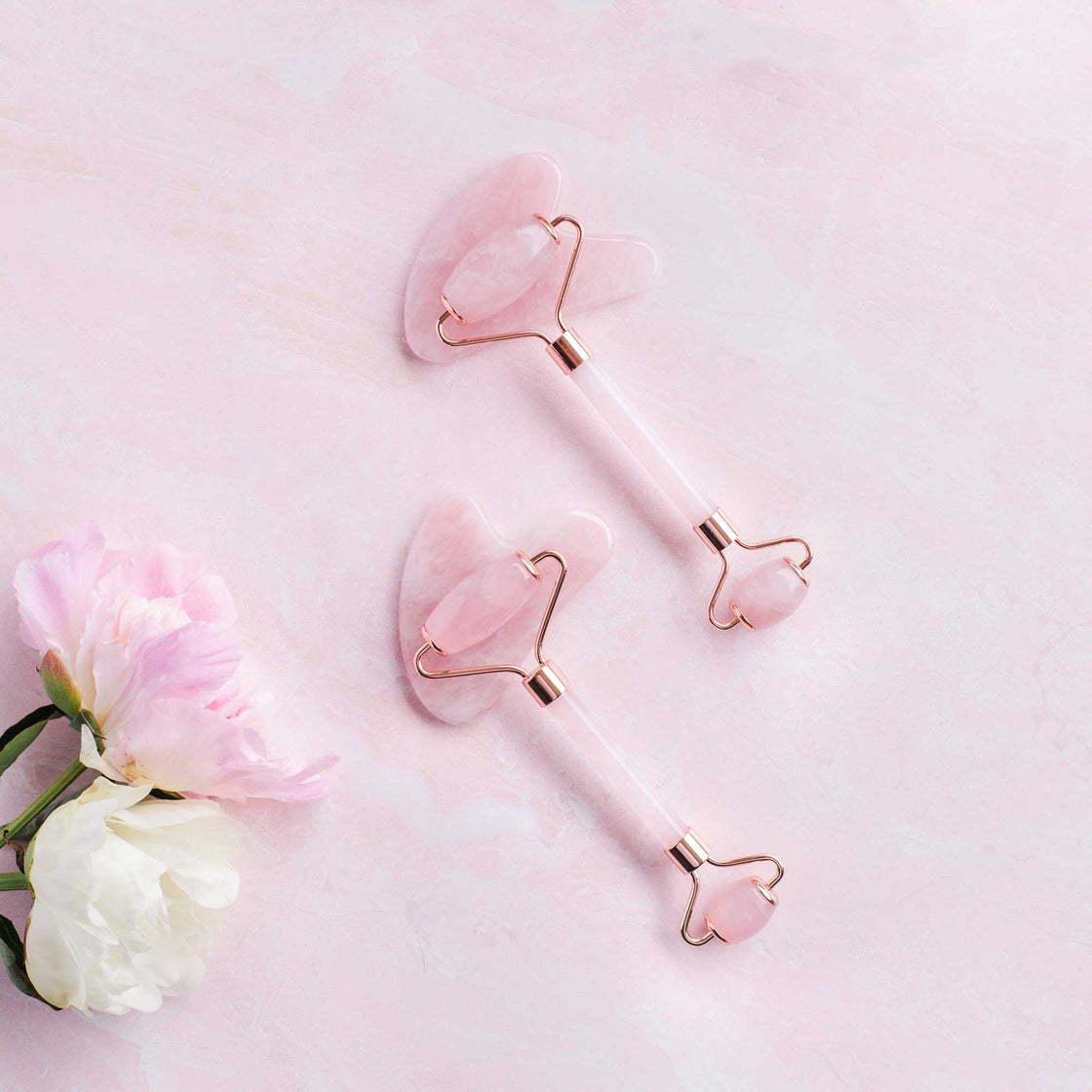 rose quartz facial rollers and gua sha set pictured on pink background with fresh pink and white peonies