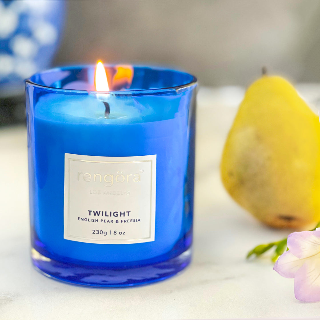 The rengöra twilight scented home candle is captured in the moment of being lit, with the fragrant notes of English pear and freesia, complemented by a zesty lime fruit and a delicate flower, all set against a softly blurred background.
