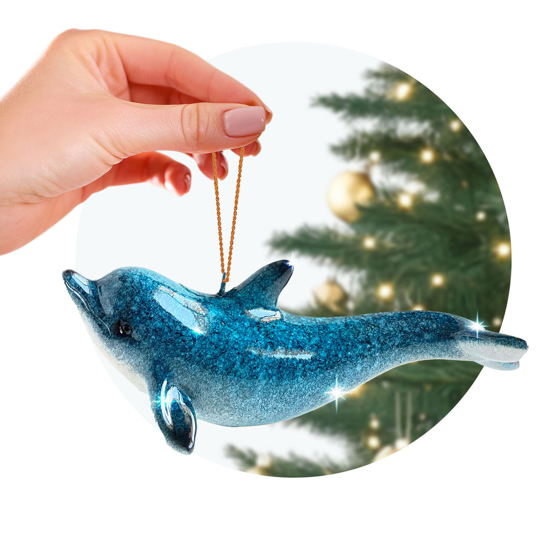 A person gently lifts the blue dolphin ornament by its gold loop, set against a festive backdrop with golden baubles on a Christmas tree.