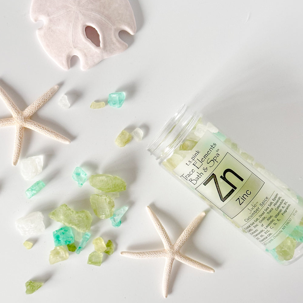 Jar of Rengöra colorful bath salts, in shades of green, white, and turquoise, are scattered around the bottle. Starfish and a sand dollar decorate the scene, enhancing a coastal, spa-like aesthetic.