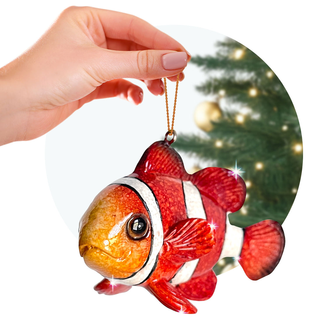 A person holds a glossy orange and white clownfish ornament by a thin gold loop, with a warmly lit Christmas tree in the background. The fish features detailed fins and a shimmering surface.