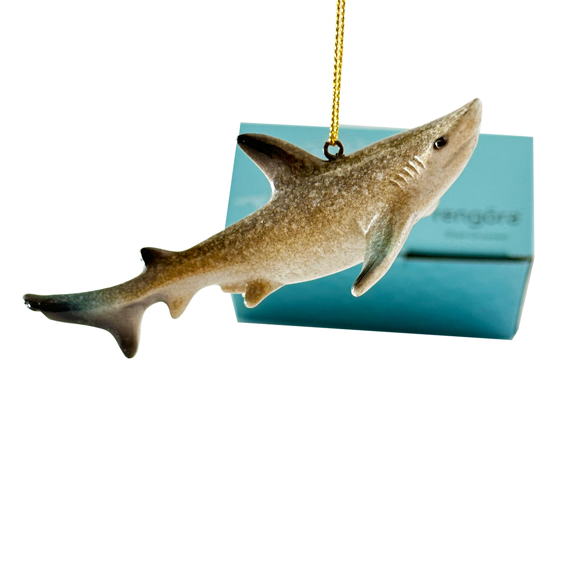 A rengöra shark ornament dangles from a Christmas tree, seen from a side perspective, with its dedicated packaging in the background against a softly blurred plain white backdrop