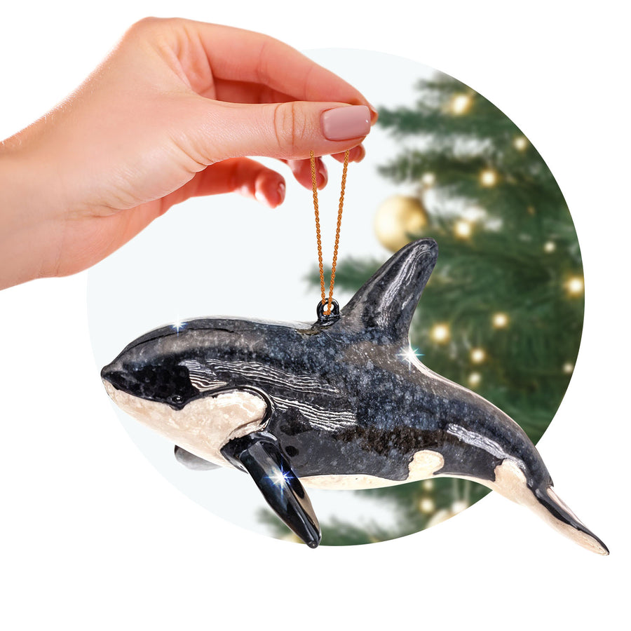 A person gently holds a black and white orca ornament by its gold loop against a festive backdrop. 