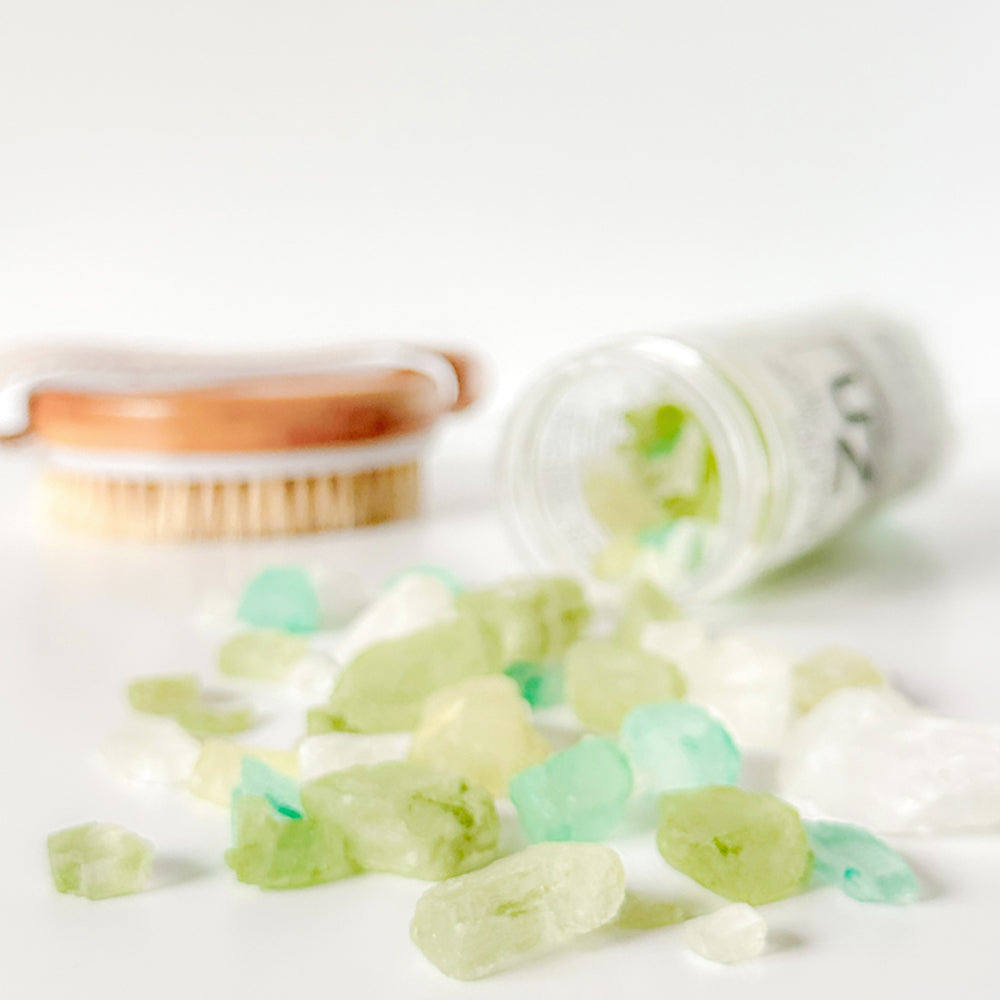 A tipped-over jar of Rengöra colorful bath salts in shades of green, white, and turquoise spills onto a white surface. 