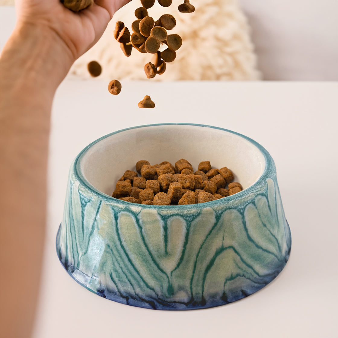 A close-up of a handcrafted blue and teal ceramic dog bowl with a unique glaze design, as food for dog is being poured into it, placed on a white table with a cozy background.