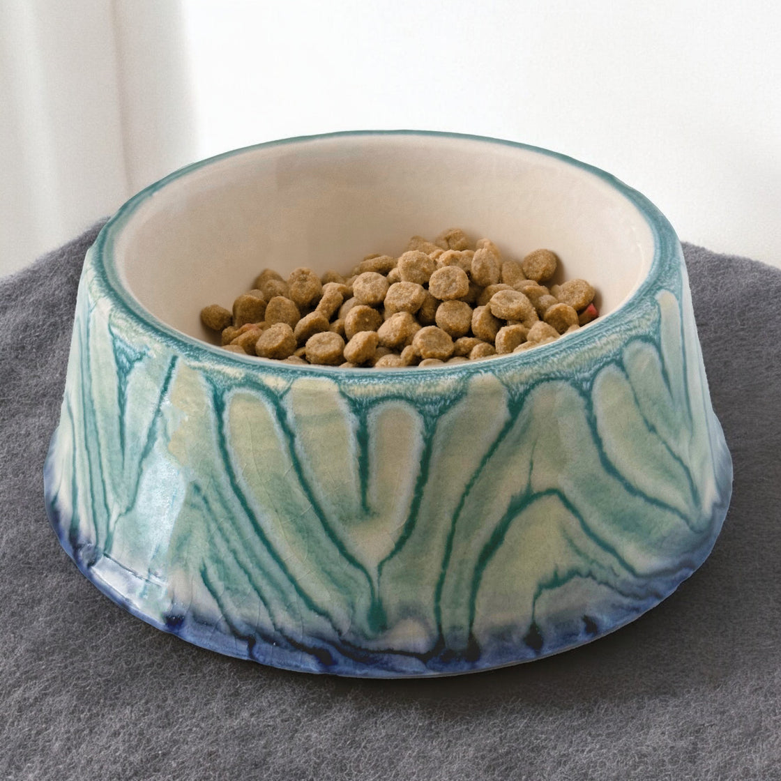 A beautifully handcrafted blue ceramic dog bowl from rengöra with a marbled teal glaze, filled with dry food, placed on a soft gray fabric surface for a cozy look.