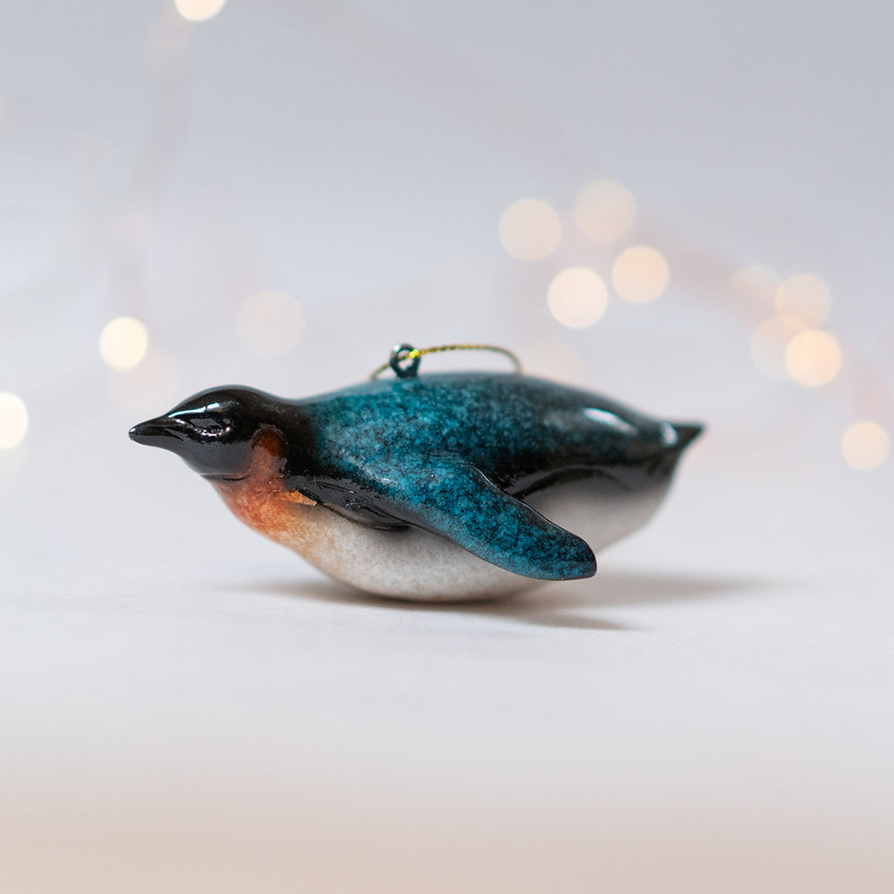 A side perspective of a Rengora penguin ornament set against a backdrop of softly blurred Christmas lights