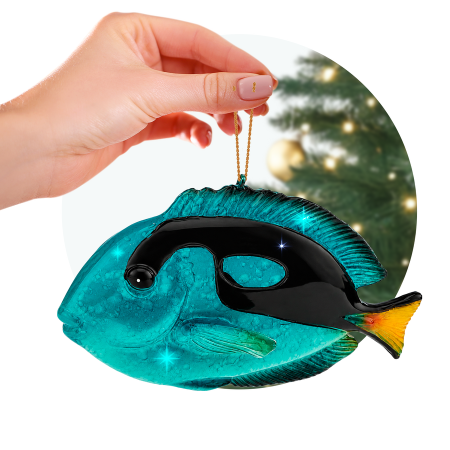 Hand holding a Rengöra tropical blue tang Christmas ornament in front of a softly lit holiday tree.