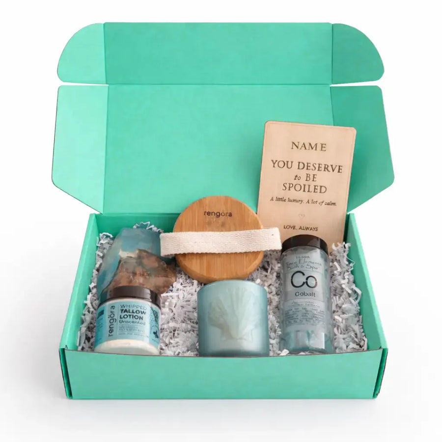 Open turquoise self care gift box with whipped tallow lotion, bath salts, soap rock, bamboo body brush, tea light holder, and engraved wooden greeting card on white background.