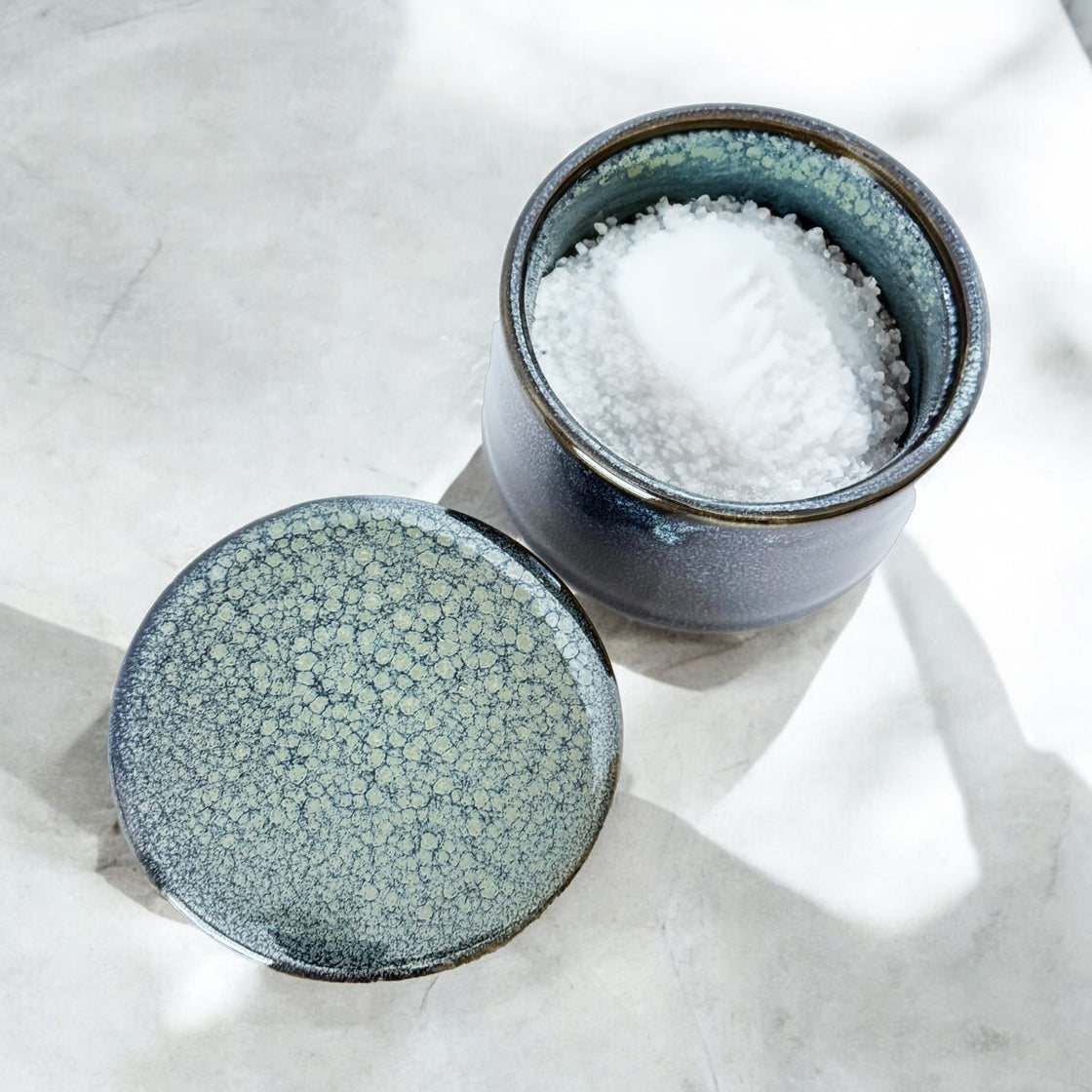 Navy ceramic storage dish with lid opened, filled with salt, showing beautiful speckled glaze on marble background.