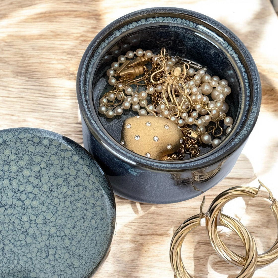 Navy ceramic covered dish holding delicate gold jewelry and pearl necklaces, placed on a wooden surface.