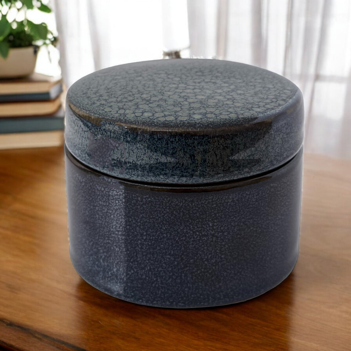 Handmade navy ceramic container with lid placed on a wooden table, next to books and houseplants, enhancing home office decor.