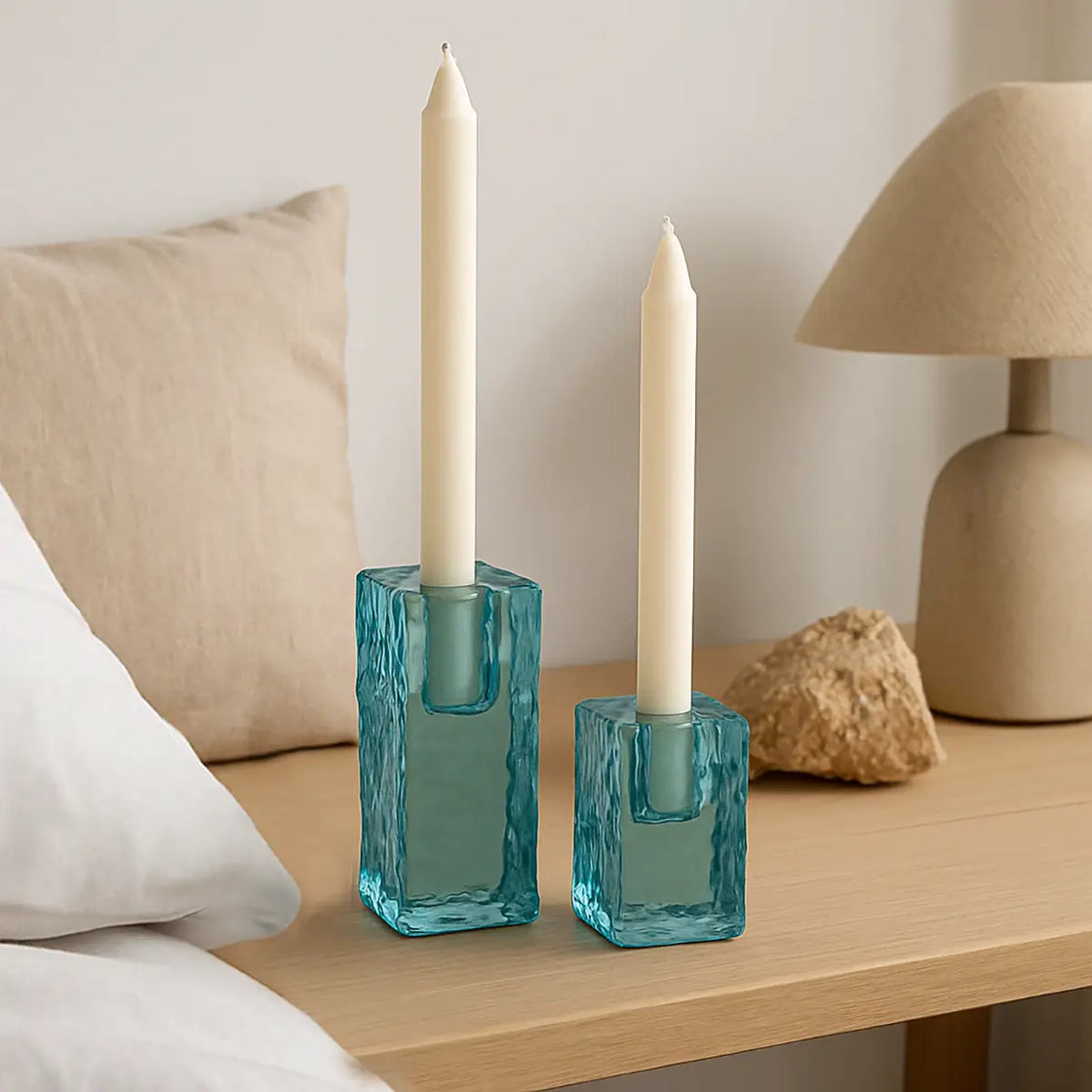 Two teal glass candle holders with white candles on a wooden surface, with a lamp and rock in the background.