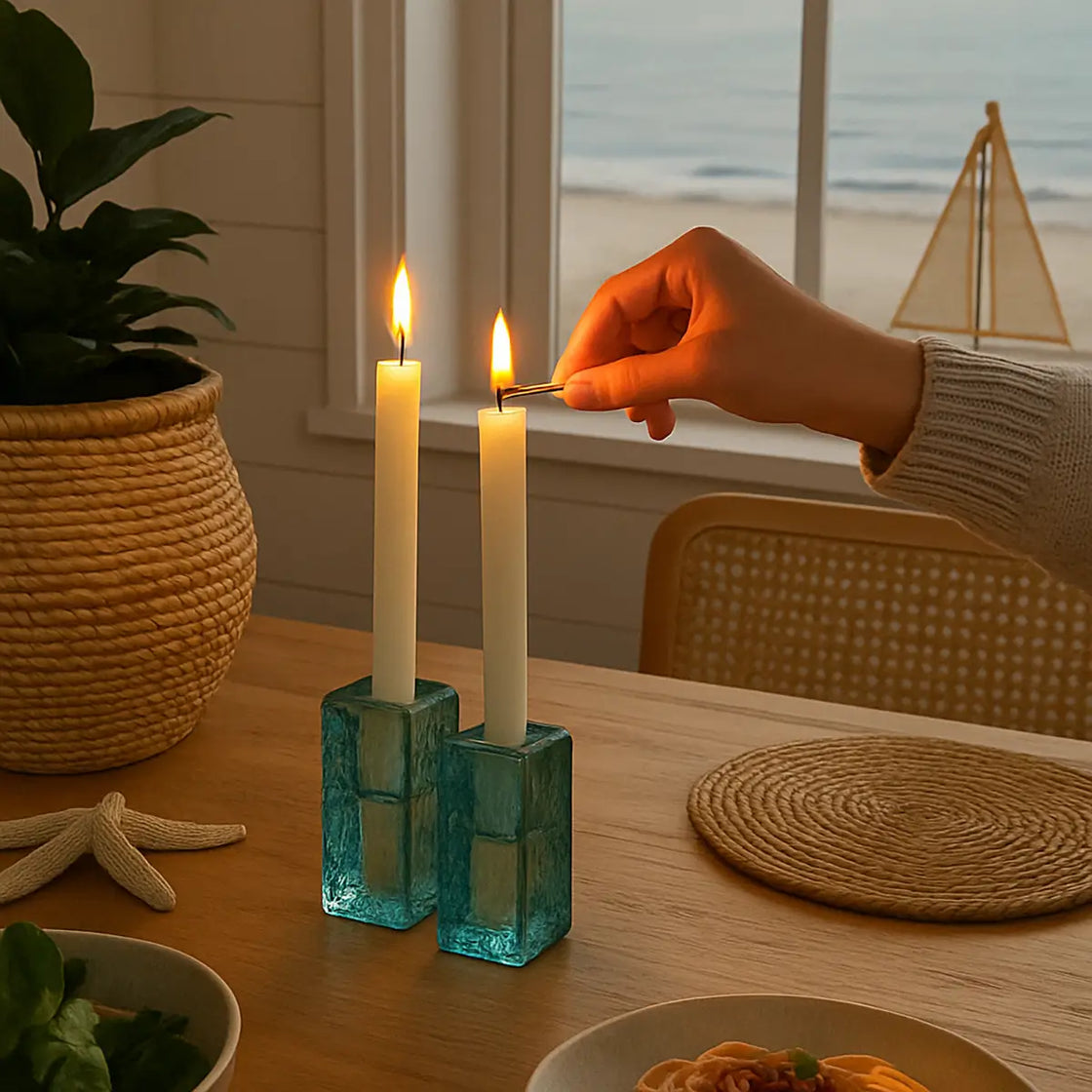 Person lighting two candles in teal holders on a table with a beach view in the background.