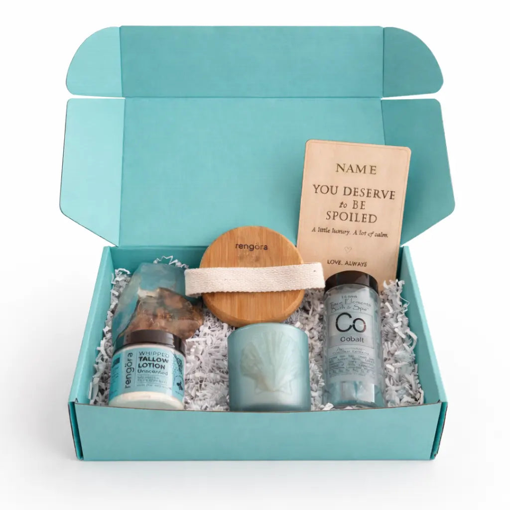 Open turquoise self care gift box with whipped tallow lotion, bath salts, soap rock, bamboo body brush, tea light holder, and engraved wooden greeting card on white background.