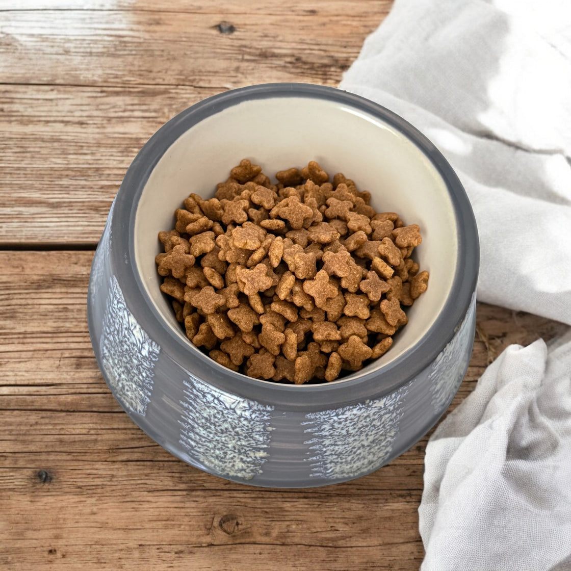 A top-down view of a gray ceramic dog bowl with a detailed marbled glaze pattern, filled with food, set on a rustic wooden table with a soft linen napkin beside it.