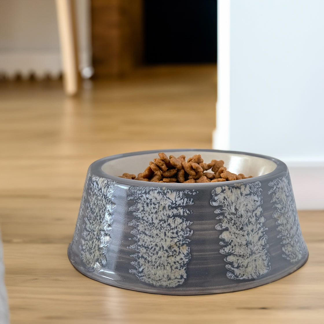 A black and white dog lying on a beige rug, gazing at a gray ceramic dog bowl filled with food, showcasing the bowl's elegant and functional design.