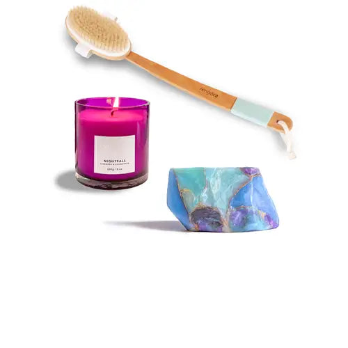 Lavender candle in a purple glass container, wooden brush, and decorative soap made to look like a gemstone (opal) on a white background