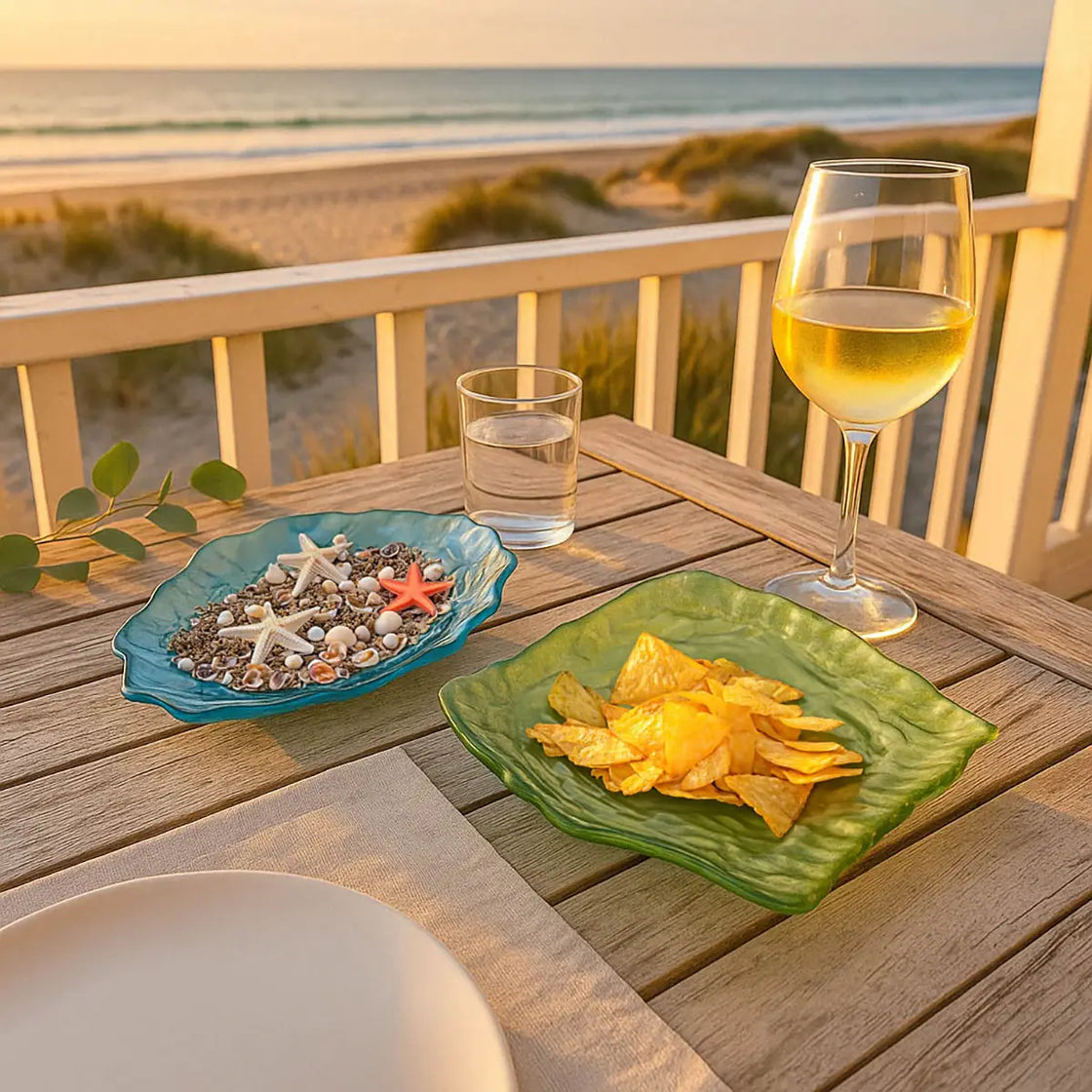 Coastal dining setup with blue and green glass oyster dishes filled with almonds and chocolates, set on a beachside deck at sunset.