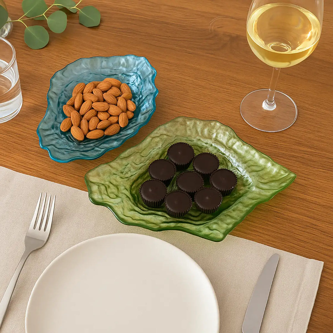 Textured glass oyster dishes used as serving bowls for almonds and chocolates on a wooden dinner table with wine and tableware.