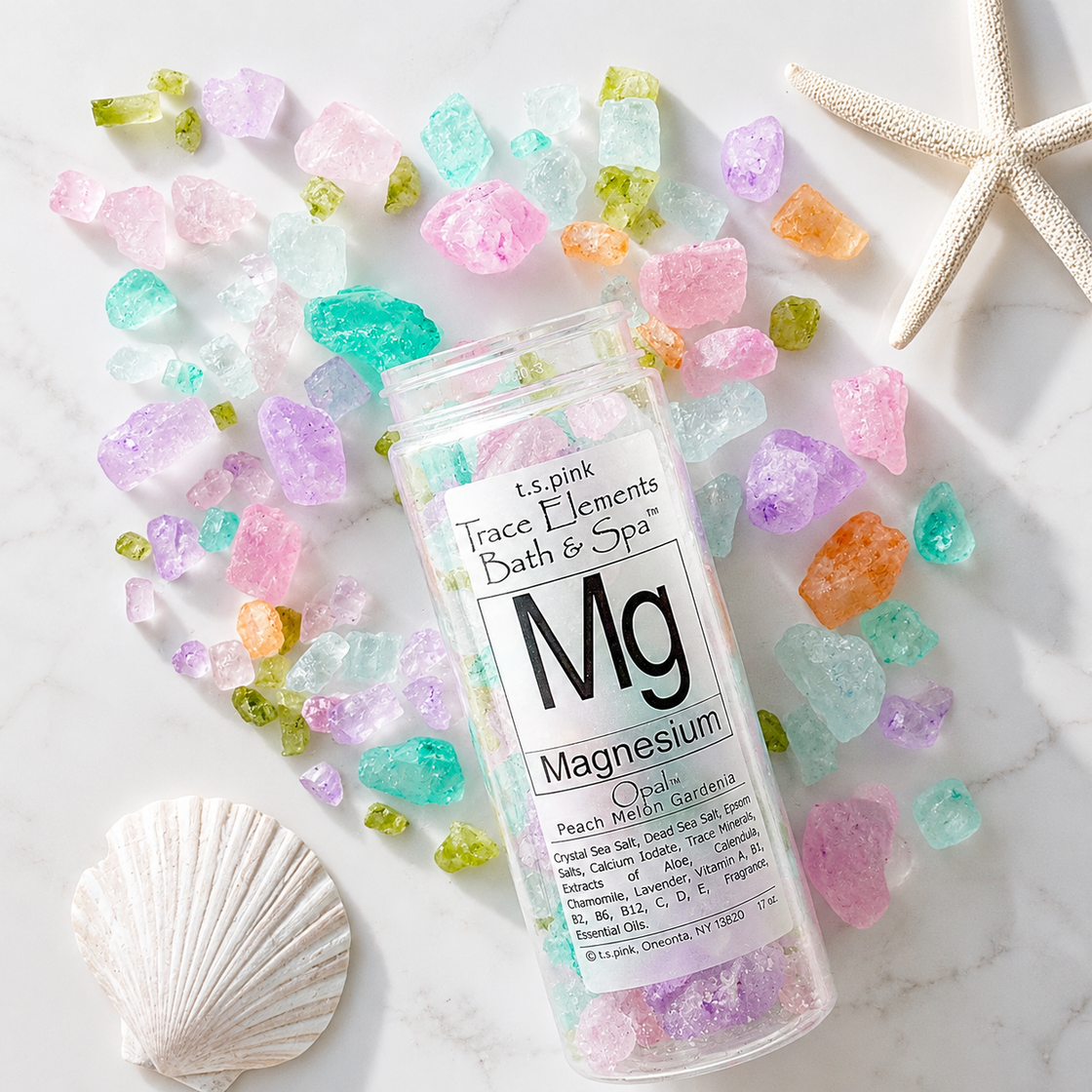 Bottle of 'Mg' magnesium bath crystals surrounded by some of thec olorful bath salts/crystals and shells on a marble surface