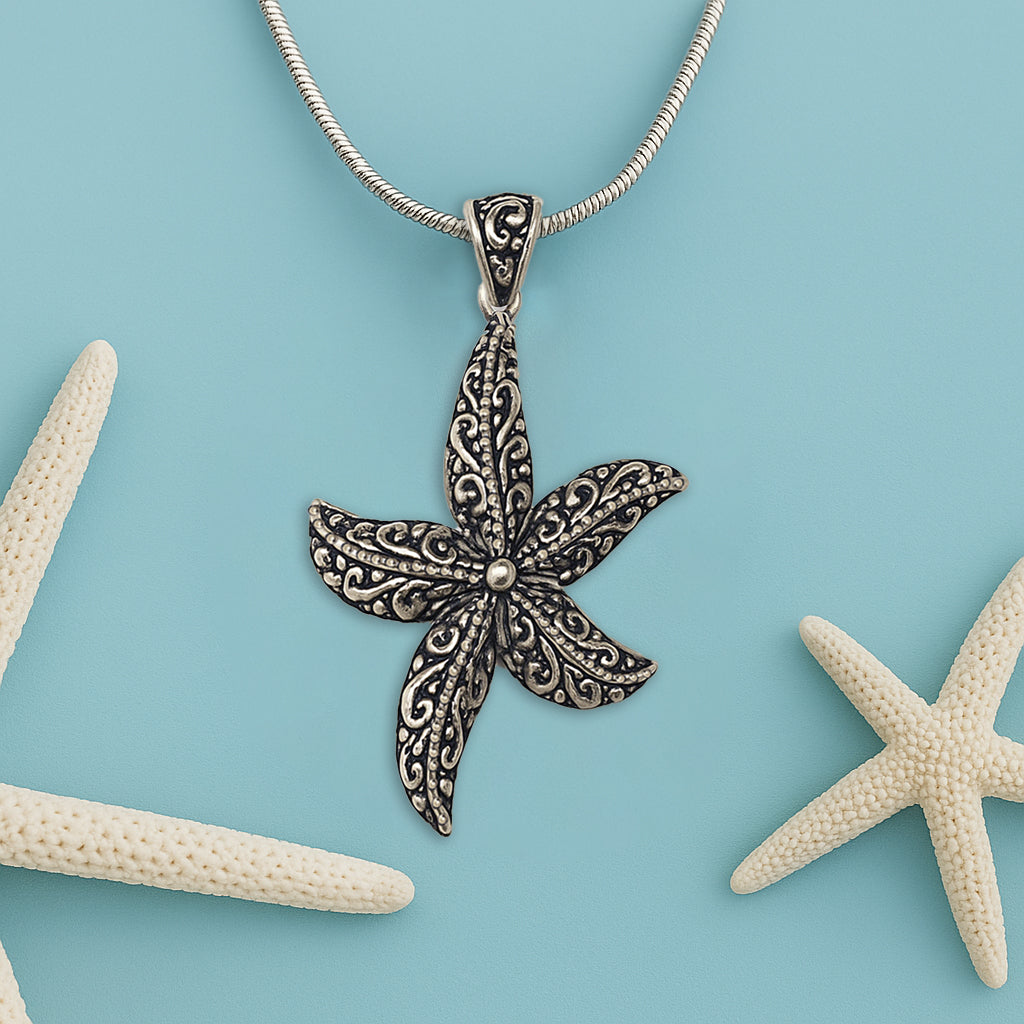 A silver starfish pendant with decorative filigree patterns displayed on a blue background, styled with two natural white starfish for a summery, beach-inspired aesthetic.