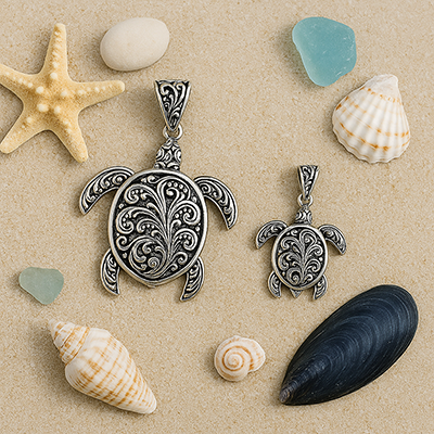 Large and small sterling silver turtle pendants placed on sand surrounded by seashells and sea glass, showcasing an ocean-inspired theme.