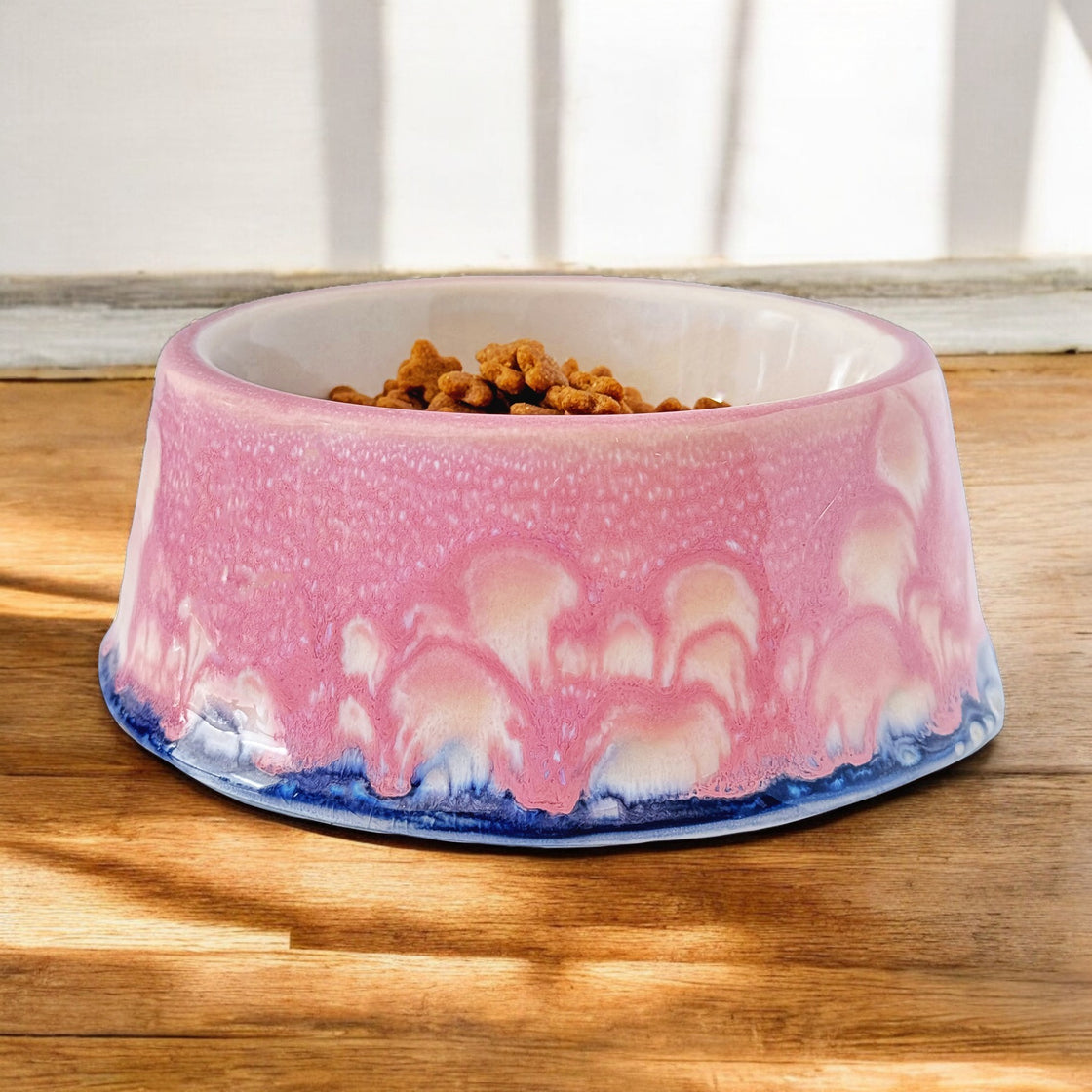 A handcrafted pink rengöra ceramic dog bowl with a unique glazed design in pink, white, and blue tones, filled with dry dog food, placed on a wooden surface with natural light.