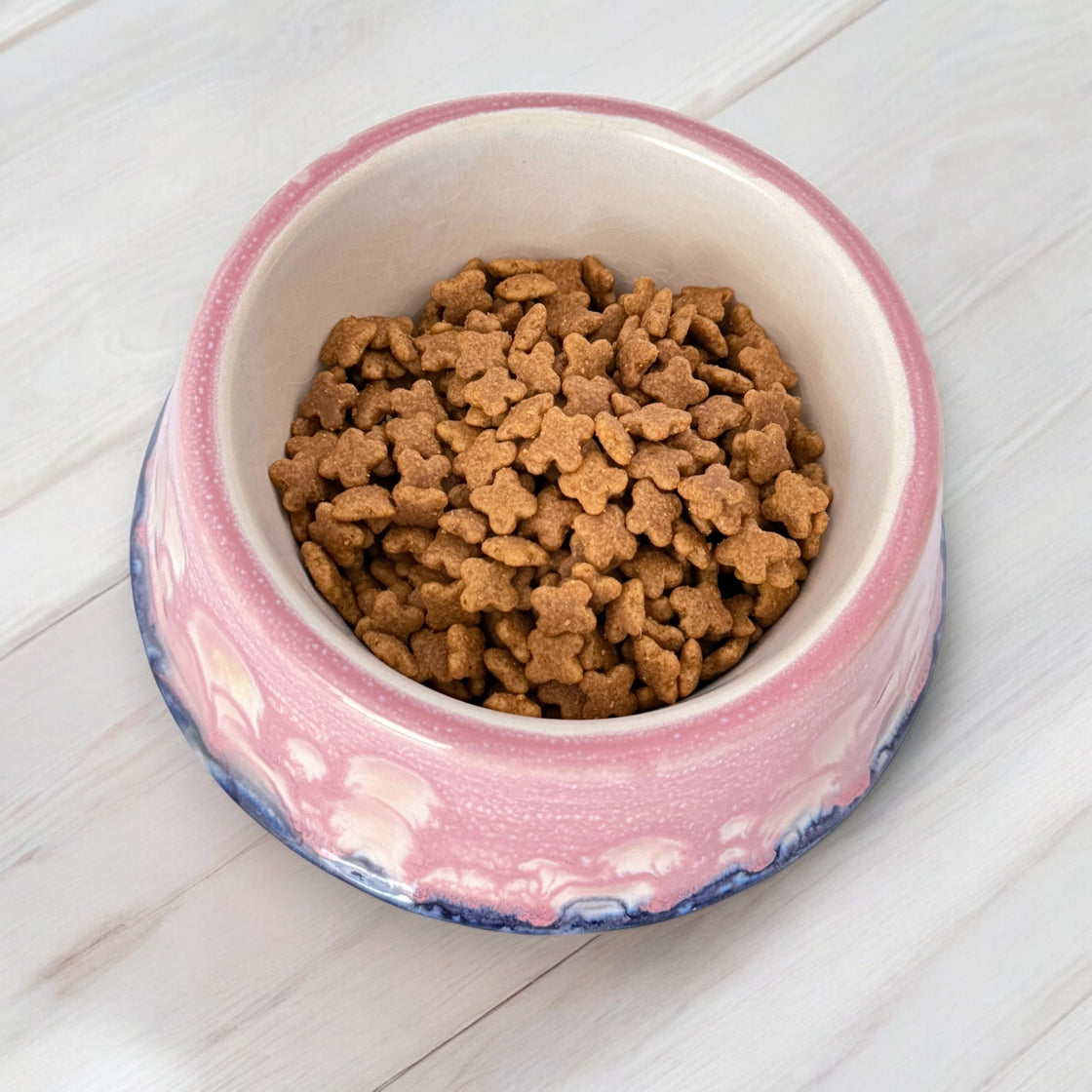 A top-down view of a pink ceramic dog bowl with a marbled pink and blue glaze, filled with small, bone-shaped dog biscuits, placed on a light wooden surface.