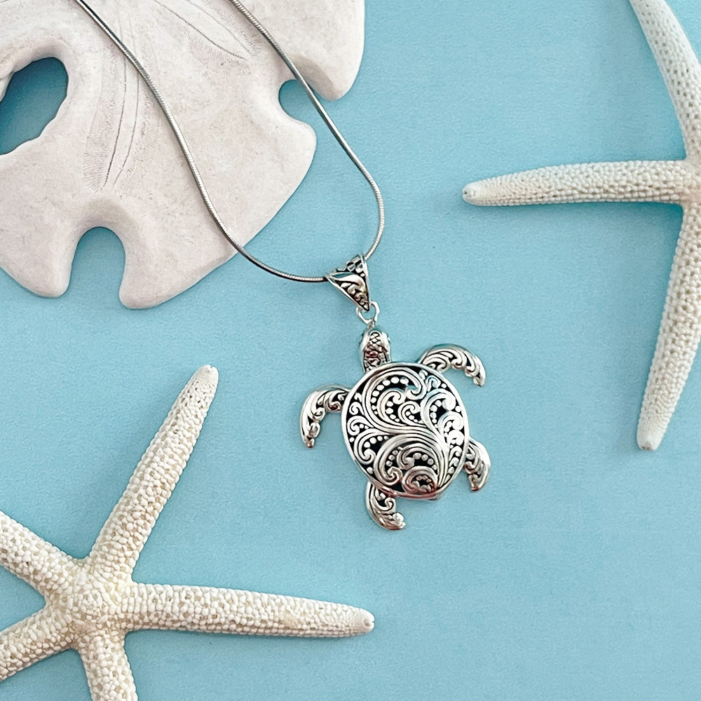 A sterling silver sea turtle pendant with intricate filigree design presented against a blue background with sea stars and a sand dollar.