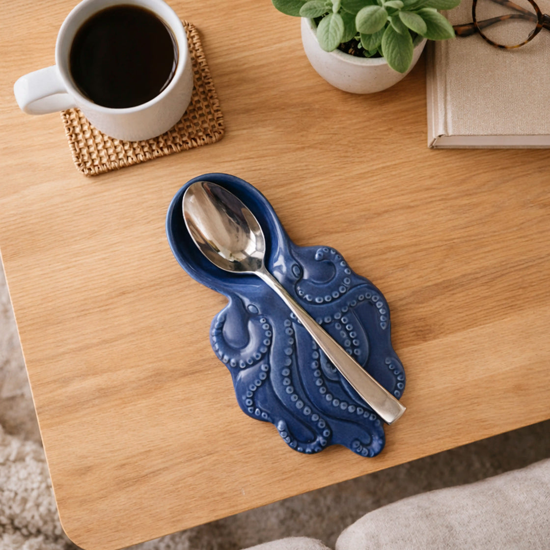 Blue decorative spoon rest with a spoon on a wooden table with a cup of coffee and plant in the background.