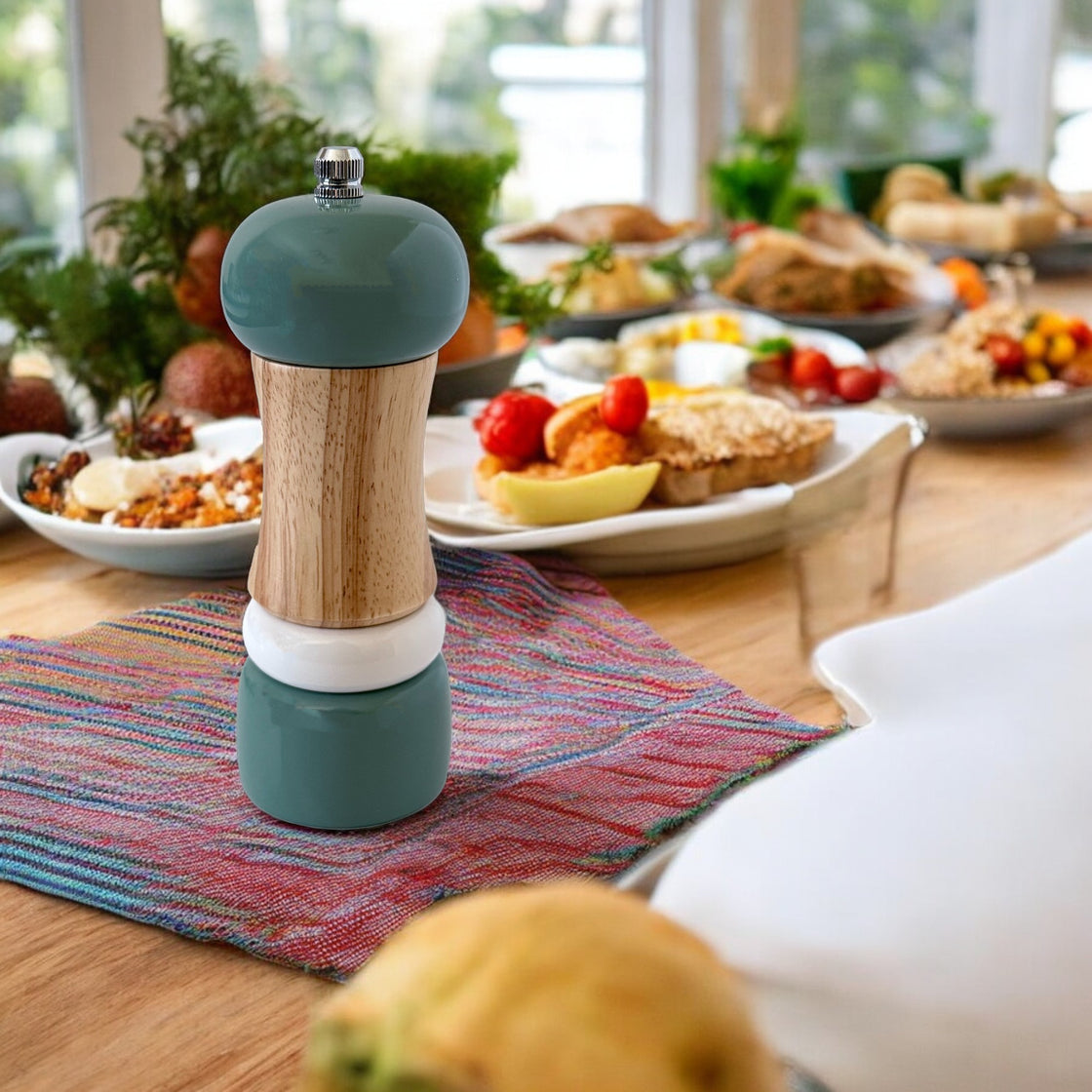 Teal wooden pepper grinder on a colorful cloth napkin, placed on a beautifully set table filled with dishes of fresh food.