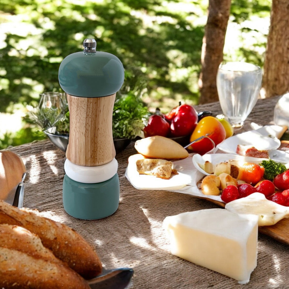 Teal wooden pepper grinder displayed elegantly on a rustic outdoor dining table, surrounded by fresh fruits, cheeses, bread, and vibrant greenery.