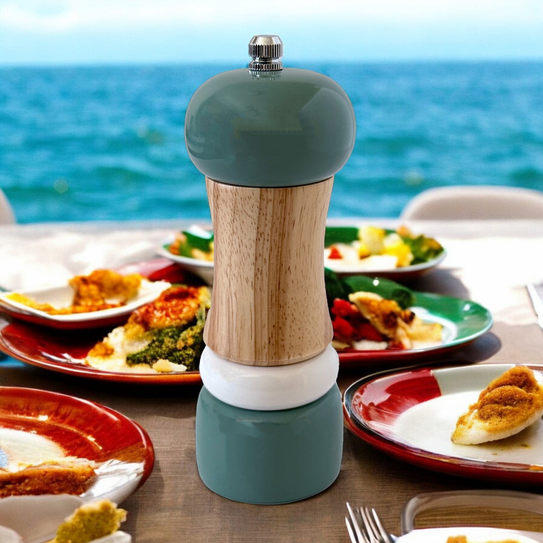 Teal wooden pepper grinder placed on an inviting seaside table setting, surrounded by delicious dishes and overlooking a tranquil ocean backdrop.