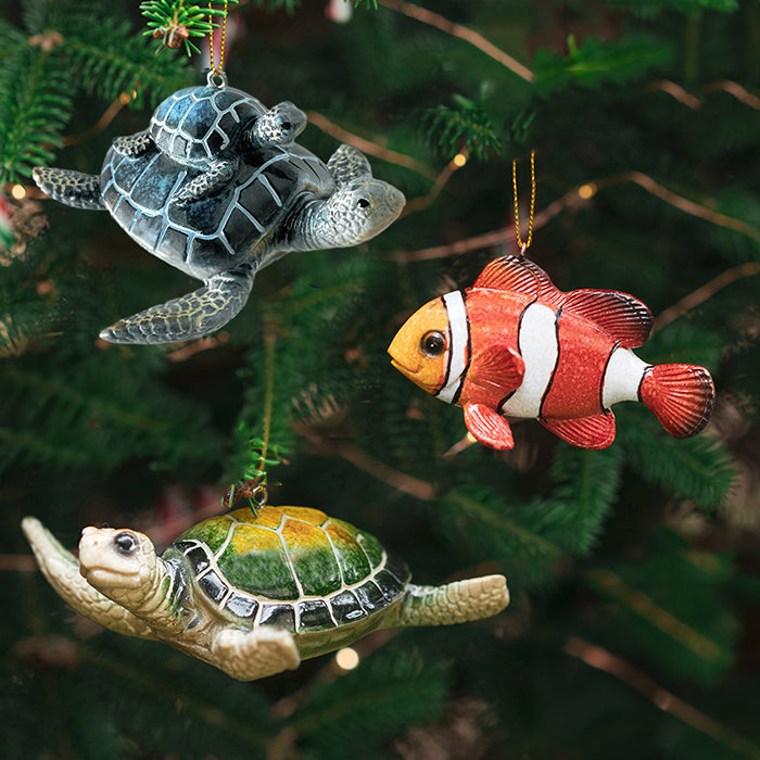 Three Christmas ornaments designed as a clownfish, a green turtle, and a mother turtle with a child turtle on her back, hanging from a tree with a blurred green background.