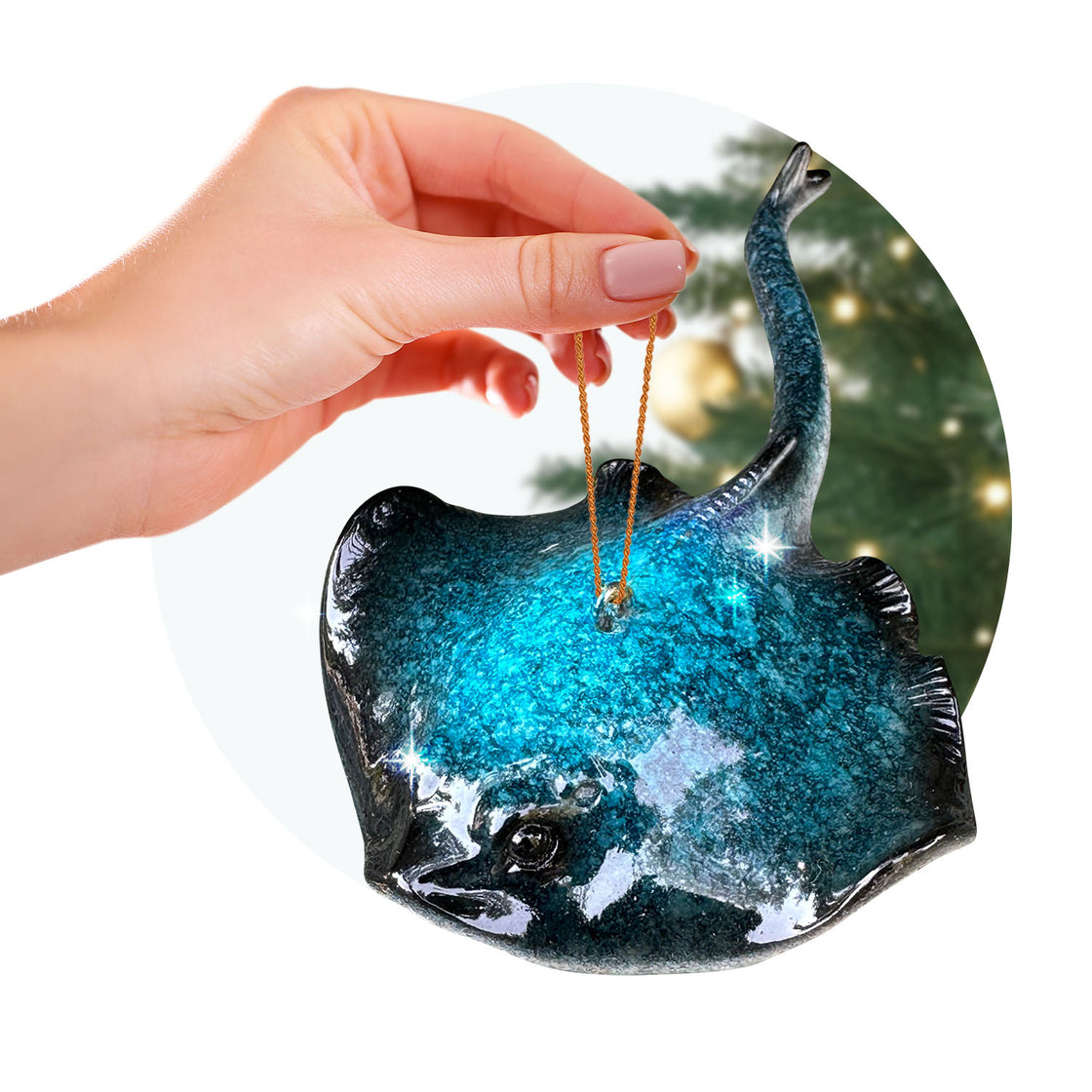 A close-up of a person holding a metallic teal stingray ornament by its gold loop, with a softly lit Christmas tree in the background. The ornament features a curved tail and glimmering finish.