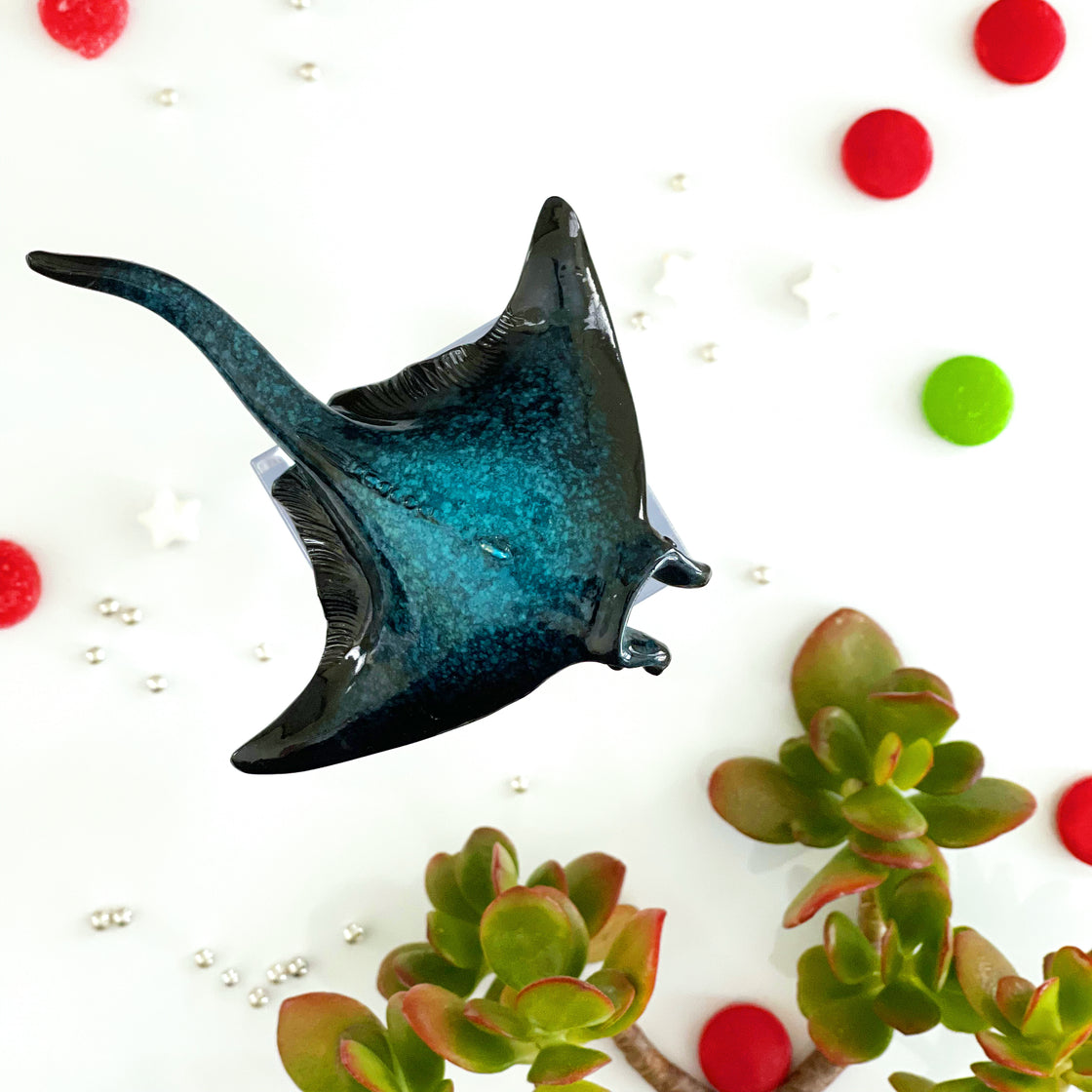A top-down perspective of the rengora manta ray Christmas ornament set against a backdrop of a green jade leaves and Christmas balls as part of the decorative arrangement