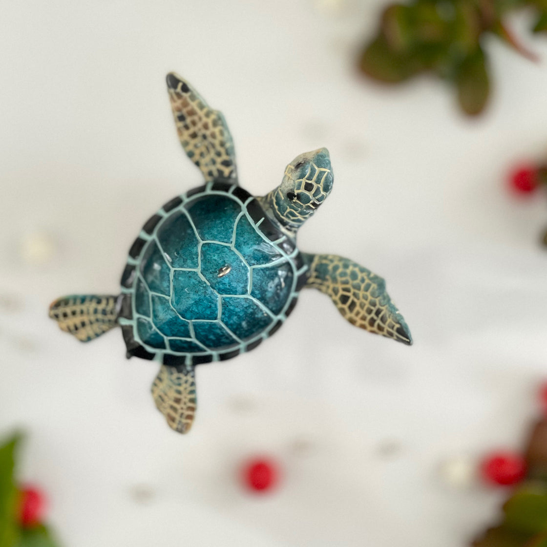 top view of blue sea turtle ornament looking like it's hovering or swimming between some green plants with red berries